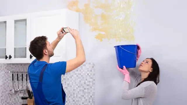 Homeowner photographing a water stained wall while a second person holds a bucket, documenting damage for a claim